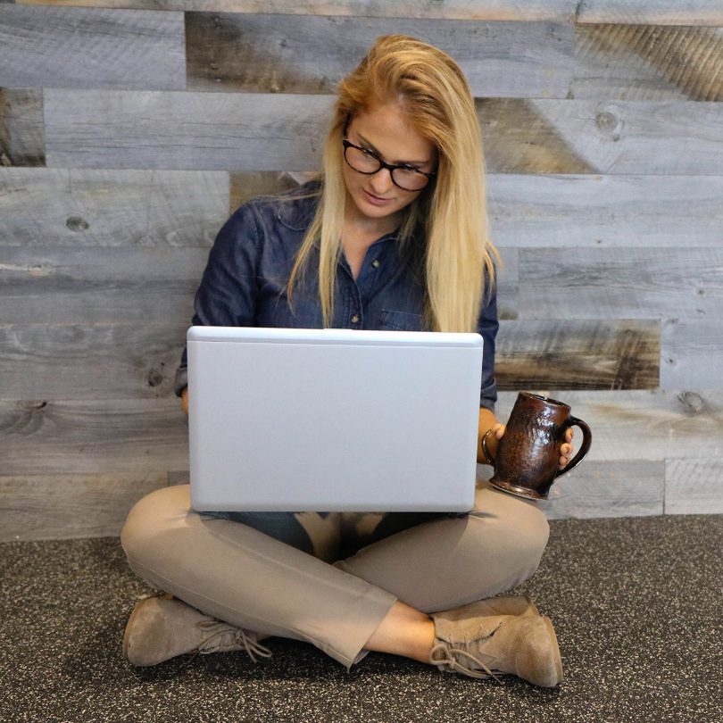 Blonde woman in glasses wearing business casual" slacks and button-down blouse sitting on the floor reading from her laptop